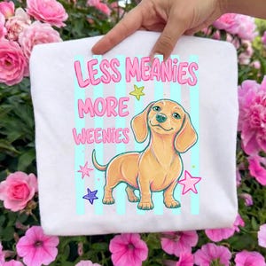 May include: White tote bag featuring a cartoon dachshund with the text "LESS MEANIES MORE WEENIES" in pink. The dog is light brown with a happy expression, set against a blue and white striped background with star accents.