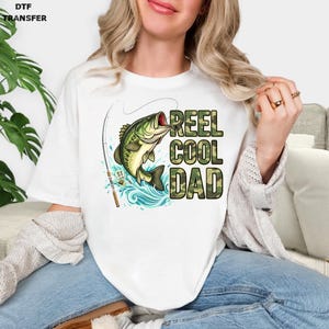 May include: White t-shirt featuring a graphic of a largemouth bass leaping from blue water, with a fishing rod and the text "REEL COOL DAD" in camouflage lettering. DTF TRANSFER is printed in the upper left corner.