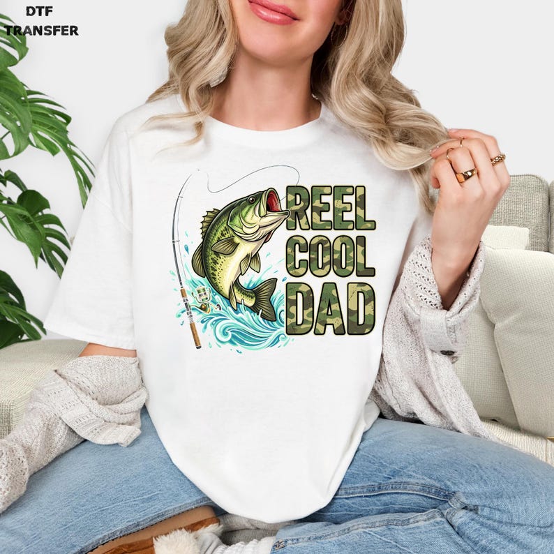 May include: White t-shirt featuring a graphic of a largemouth bass leaping from blue water, with a fishing rod and the text "REEL COOL DAD" in camouflage lettering. DTF TRANSFER is printed in the upper left corner.
