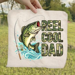 May include: A beige tote bag featuring a leaping green and black bass fish with a fishing rod. The words "REEL COOL DAD" are printed in camouflage lettering. The image is set against a green grassy background.