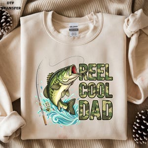 May include: Beige sweatshirt with a graphic of a green and yellow fish jumping out of blue water, with a fishing rod. The text "REEL COOL DAD" is printed in camouflage letters.