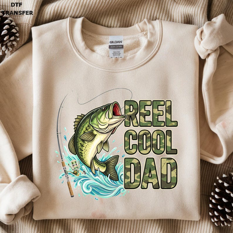 May include: Beige sweatshirt with a graphic of a green and yellow fish jumping out of blue water, with a fishing rod. The text "REEL COOL DAD" is printed in camouflage letters.