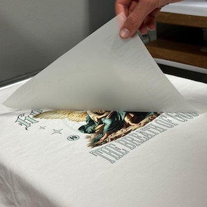 May include: White t-shirt with a graphic design featuring an angel, wings, and the text "THE BREATH OF GOD." A hand is lifting a sheet of transfer paper, revealing the design. The design includes the number "19" and star shapes.
