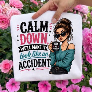 May include: A white t-shirt featuring a graphic of a person with a messy bun wearing sunglasses and holding a mug that says NOT MY PROBLEM. The text reads CALM DOWN, WE'LL MAKE IT look like an ACCIDENT in black, pink, and teal lettering.