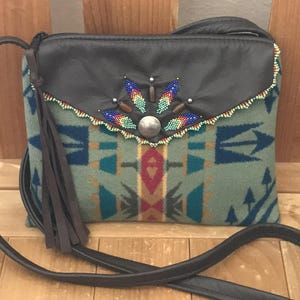 May include: A crossbody bag with a black leather flap and strap. The bag features a patterned fabric body in shades of green, blue, and red. The flap is decorated with colorful beads and a silver button. A brown tassel hangs from the side.