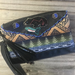May include: A black leather clutch with a wrist strap and black fringe. The flap features intricate beadwork depicting a black bear, a fish, and two paw prints. The lower section displays a patterned fabric with mountain and forest motifs.