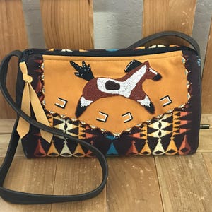 May include: A rectangular shoulder bag with a brown leather strap. The bag features a tan suede panel with a beaded horse design. The bag's body has a geometric pattern in shades of brown, orange, and blue.