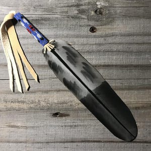 May include: A decorative feather with a beaded handle in blue, red, and white. The feather is primarily black with gray accents and has a tan leather fringe attached.