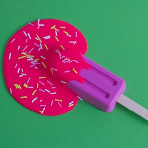 Puede incluir: Una paleta de hielo rosa con un palo blanco, derriti&eacute;ndose sobre una superficie verde. La paleta est&aacute; cubierta de chispas rosas y blancas.