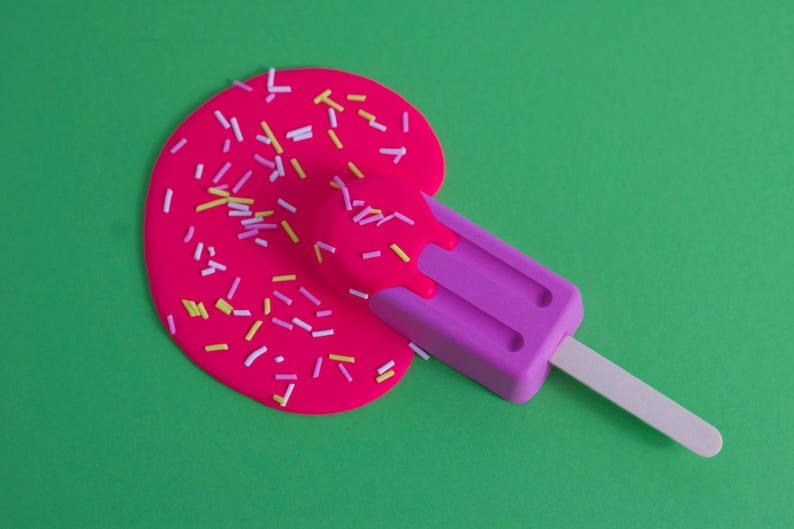 Puede incluir: Una paleta de hielo rosa con un palo blanco, derriti&eacute;ndose sobre una superficie verde. La paleta est&aacute; cubierta de chispas rosas y blancas.