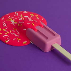 May include: A pink popsicle with a wooden stick is melting on a purple surface. The popsicle is covered in pink and white sprinkles.