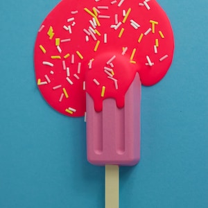 Puede incluir: Una paleta de hielo rosa con un palo de madera se derrite sobre un fondo azul. La paleta est&aacute; cubierta de glaseado rosa y chispas blancas y amarillas.