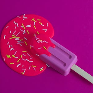 Puede incluir: Una paleta de hielo rosa con un palo de madera se derrite sobre un fondo rosa con chispas rosas y blancas.