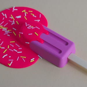 Puede incluir: Una paleta de hielo rosa con un palo blanco, derriti&eacute;ndose sobre una superficie rosa cubierta de chispas de colores.