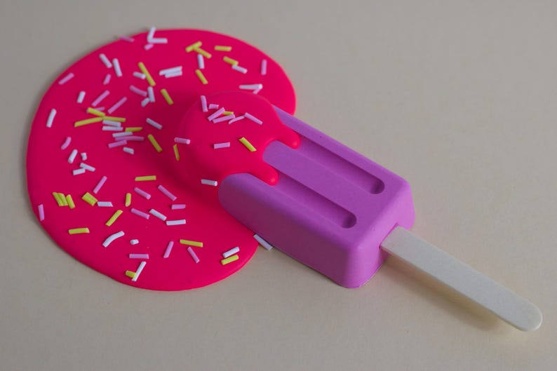Puede incluir: Una paleta de hielo rosa con un palo blanco, derriti&eacute;ndose sobre una superficie rosa cubierta de chispas de colores.