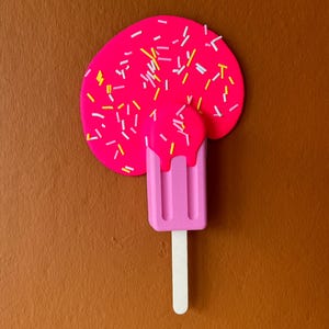 Puede incluir: Una paleta de hielo rosa con chispas blancas derriti&eacute;ndose sobre una superficie marr&oacute;n. La paleta de hielo tiene tres crestas y un palo blanco.