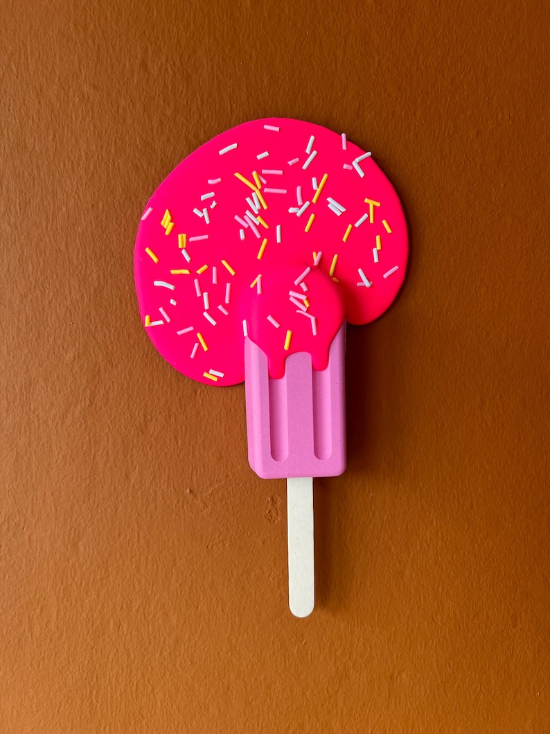 Puede incluir: Una paleta de hielo rosa con chispas blancas derriti&eacute;ndose sobre una superficie marr&oacute;n. La paleta de hielo tiene tres crestas y un palo blanco.