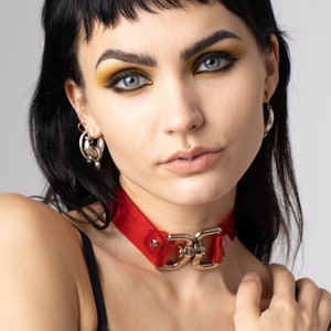 Puede incluir: Una mujer con el pelo oscuro y un maquillaje de ojos llamativo lleva un collar rojo con herrajes plateados. Lleva un top de encaje negro y tiene tatuajes en los brazos.