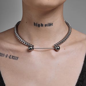 Puede incluir: Un collar choker plateado con dos grandes cuentas redondas plateadas conectadas por una barra recta. El collar lo lleva una persona con un tatuaje que dice "high vibe" en el cuello.