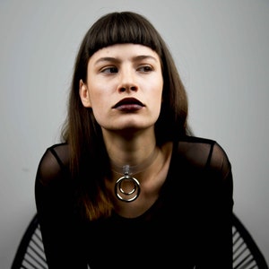Könnte beinhalten: Eine Frau mit dunkelbraunen Haaren und Pony trägt ein schwarzes, durchsichtiges Oberteil mit einer durchsichtigen Halskette mit einem silbernen Doppelring-Anhänger.