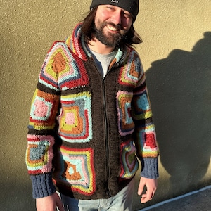 Puede incluir: Un hombre con una boina negra con la palabra "Do" bordada, un suéter de crochet marrón con cuadrados coloridos y pantalones vaqueros azules.