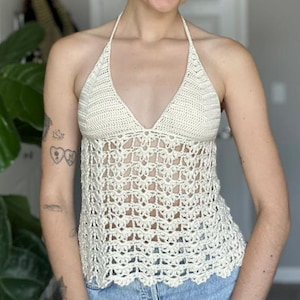 May include: Cream-colored crochet halter top with a V-neck design. The top features an open, lacy pattern and a scalloped hemline. The top is paired with blue denim jeans.