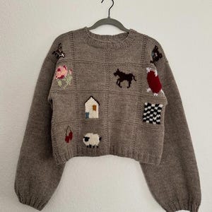 Op de afbeelding: Een taupekleurige gebreide trui met een ruitpatroon en diverse intarsia-motieven, waaronder een roze bloem, een donker paard, een wit huis, een schaap, kersen, een rode vorm en een zwart-wit geblokt vierkant, met lange mouwen en een ronde hals.