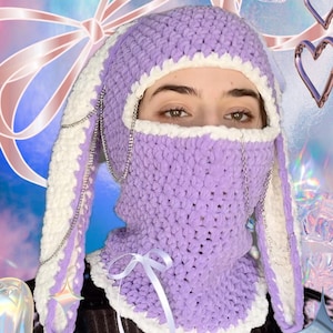 May include: A purple crocheted balaclava with long bunny ear-shaped flaps, trimmed in white and featuring a white ribbon tied at the neck. The balaclava has a chain detail around the face.