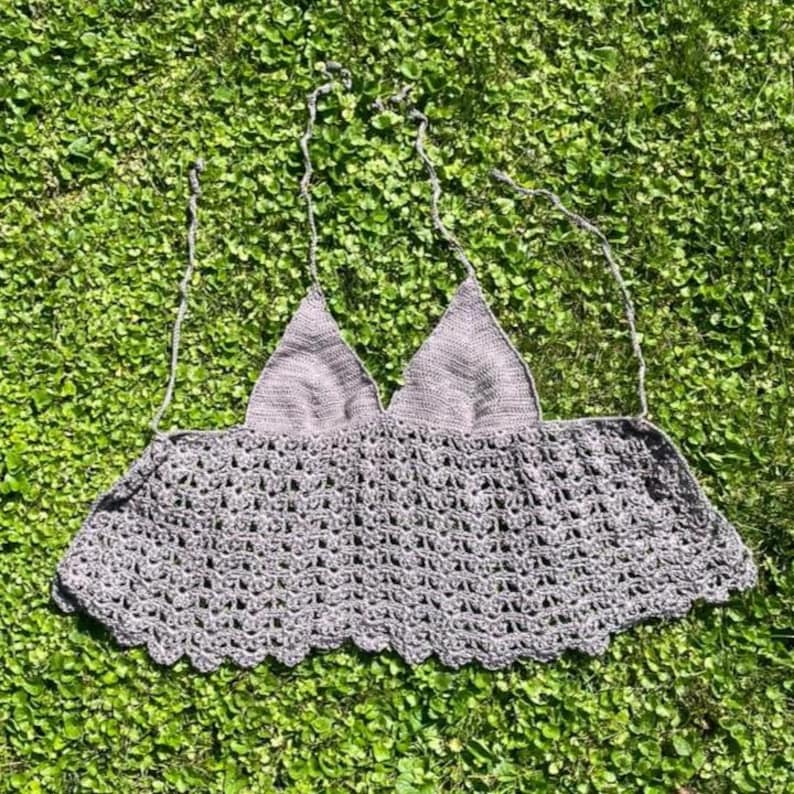 May include: A gray crochet top with a halter neck design. The top features a triangle-shaped bust and a scalloped hemline. The open weave pattern is visible throughout the garment, which is lying on a bed of green grass.