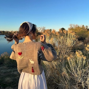 May include: A beige knit sweater featuring a grid pattern with embroidered motifs including a red tulip, a white rabbit, a red lighthouse, and a red strawberry. The sweater has long sleeves and a crew neckline, paired with a long white skirt.