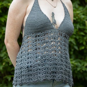 May include: A gray halter top with a crochet design. The top features a halter neck and a detailed openwork pattern across the chest and torso. The bottom hem has a scalloped edge. The top is paired with light blue denim jeans.