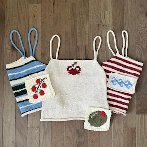 May include: Three knitted tank tops with spaghetti straps. The left top has blue and green stripes with a square patch featuring red strawberries. The center top is cream with a red crab motif and a square patch showing a green olive. The right top has red and white stripes with a square patch featuring blue waves.