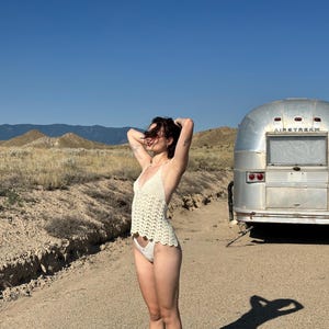 May include: A cream-colored crochet top and matching bottoms. A vintage silver Airstream trailer is parked on a dirt road. The background features dry grass, mountains, and a clear blue sky.