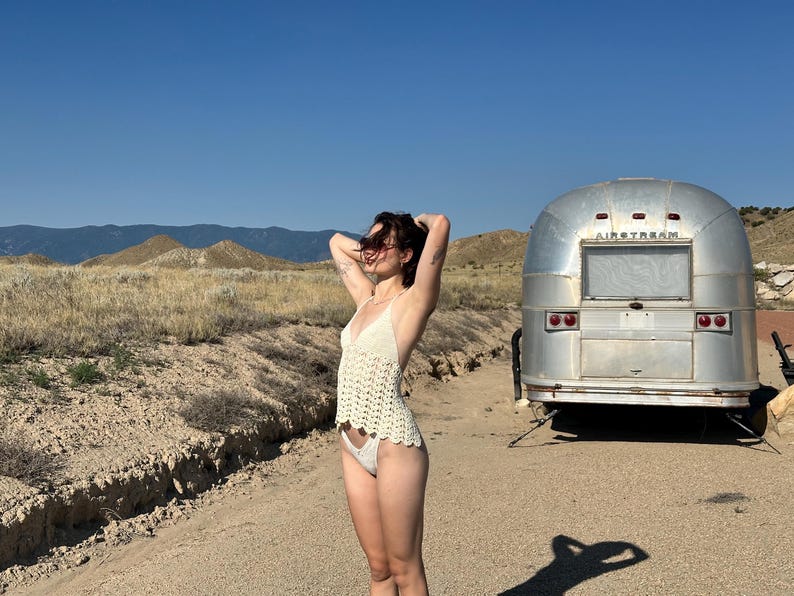 May include: A cream-colored crochet top and matching bottoms. A vintage silver Airstream trailer is parked on a dirt road. The background features dry grass, mountains, and a clear blue sky.