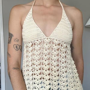 May include: Cream-colored crochet halter top with a deep V-neck and openwork design. The top features a delicate floral pattern and a scalloped hemline, offering a bohemian aesthetic. The halter straps are thin and the top is made of a soft, textured yarn.