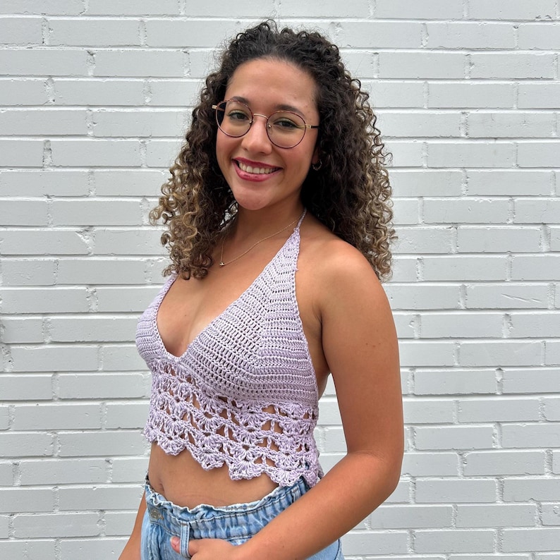 May include: A light purple crochet halter top with a scalloped hem. The top is made of a soft, textured yarn and features a deep V-neckline. The top is paired with blue denim jeans.
