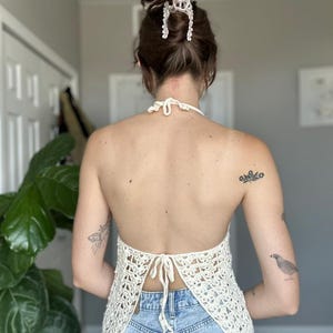 May include: Cream-colored, open-back crochet top with a halter neck and tie closure. The top is made of a delicate, open-weave pattern. The model is wearing blue jeans. A white hair clip is in the model's hair.