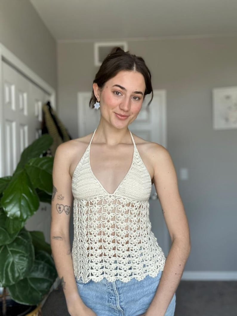 May include: Cream-colored crochet halter top with a V-neck design. The top features an open, lacy pattern and a scalloped hemline. The top is paired with blue denim jeans.