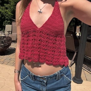 May include: A red halter top with a deep V-neck and a scalloped hem. The top is made of a crocheted material. The top is paired with blue denim jeans.