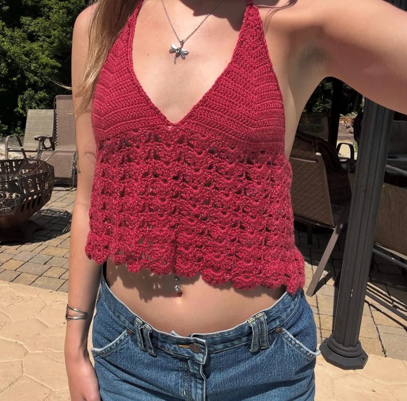 May include: A red halter top with a deep V-neck and a scalloped hem. The top is made of a crocheted material. The top is paired with blue denim jeans.