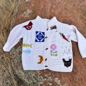 Peut inclure: Cardigan blanc en tricot avec divers patchs brodés. Les patchs incluent un oiseau rouge, un motif géométrique bleu et blanc, une carotte, une fleur et une poule noire. Le cardigan a une fermeture boutonnée et des manches longues.