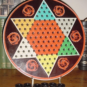 May include: A vintage Chinese Checkers board game with a star-shaped design in orange, green, yellow, and teal. The board is round with dragon motifs and comes with black and red game pieces. The game is displayed on a wooden surface.