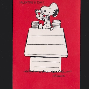 May include: A Valentine's Day card featuring Snoopy holding a heart-shaped card, sitting atop his doghouse. The card is red with white text that reads "EVERYONE AND HIS DOG WISH YOU A HAPPY VALENTINE'S DAY!" Signed "Schulz".