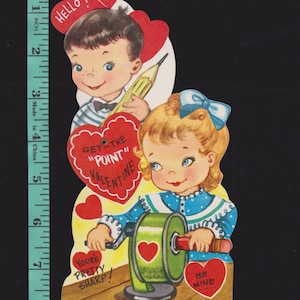 May include: Vintage Valentine's Day card featuring two children. A boy holds a pencil, and a girl sharpens one. The card has heart-shaped accents and text that reads "Hello!", "Get the Point Valentine", "You're Pretty Sharp!", and "Be Mine".