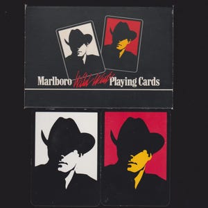 May include: A black rectangular box labeled Marlboro Wild West Playing Cards with two card illustrations featuring a cowboy silhouette. Below the box are two individual playing cards, one with a white background and one with a red background.