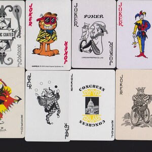 May include: A collection of nine vintage playing cards, each featuring a unique Joker illustration. The designs include a Garfield character, a jester on a bicycle, a Congress building graphic, and various colorful jester figures with text labels.