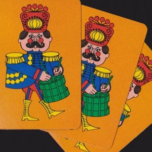 May include: Three playing cards with an orange background featuring a drummer in a blue uniform, red hat, and yellow boots. A blue measuring tape is visible on the left side, showing the card height is approximately 3.5 inches long.