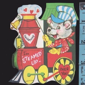 May include: Vintage Valentine's Day card featuring a cartoon bear driving a red train. The train has a heart-shaped wheel with "Over You" and a steam engine with "I'm Steamed Up" text. The card includes a bell and a "Valentine" heart.