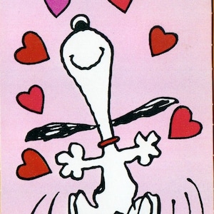 May include: Illustration of Snoopy, a white cartoon dog, jumping with joy, surrounded by red and pink hearts on a pink background. Snoopy has a red collar and is smiling. The artist's signature, Schulz, is in the bottom right corner.