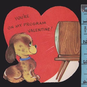 May include: Vintage Valentine's Day card featuring a brown puppy with a blue collar gazing at a wooden television set. The card is heart-shaped and has the text "YOU'RE ON MY PROGRAM VALENTINE!" The card is approximately 4 inches tall.